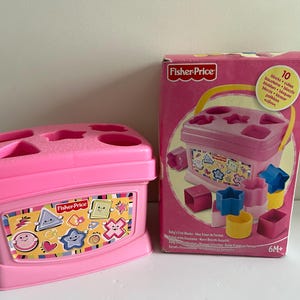 Peut inclure: Un jouet rose Fisher-Price avec un trieur de formes et sa boîte. Le trieur a des découpes de formes sur le dessus et un autocollant avec des formes et des personnages sur le devant. La boîte indique "10 blocs + cubes".