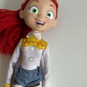 May include: A doll with bright red yarn hair, large green eyes, and an open-mouthed smile. The doll wears a white long-sleeved shirt with a yellow yoke featuring red stitching, two white buttons, blue denim shorts, and white pants with black cow-print spots.