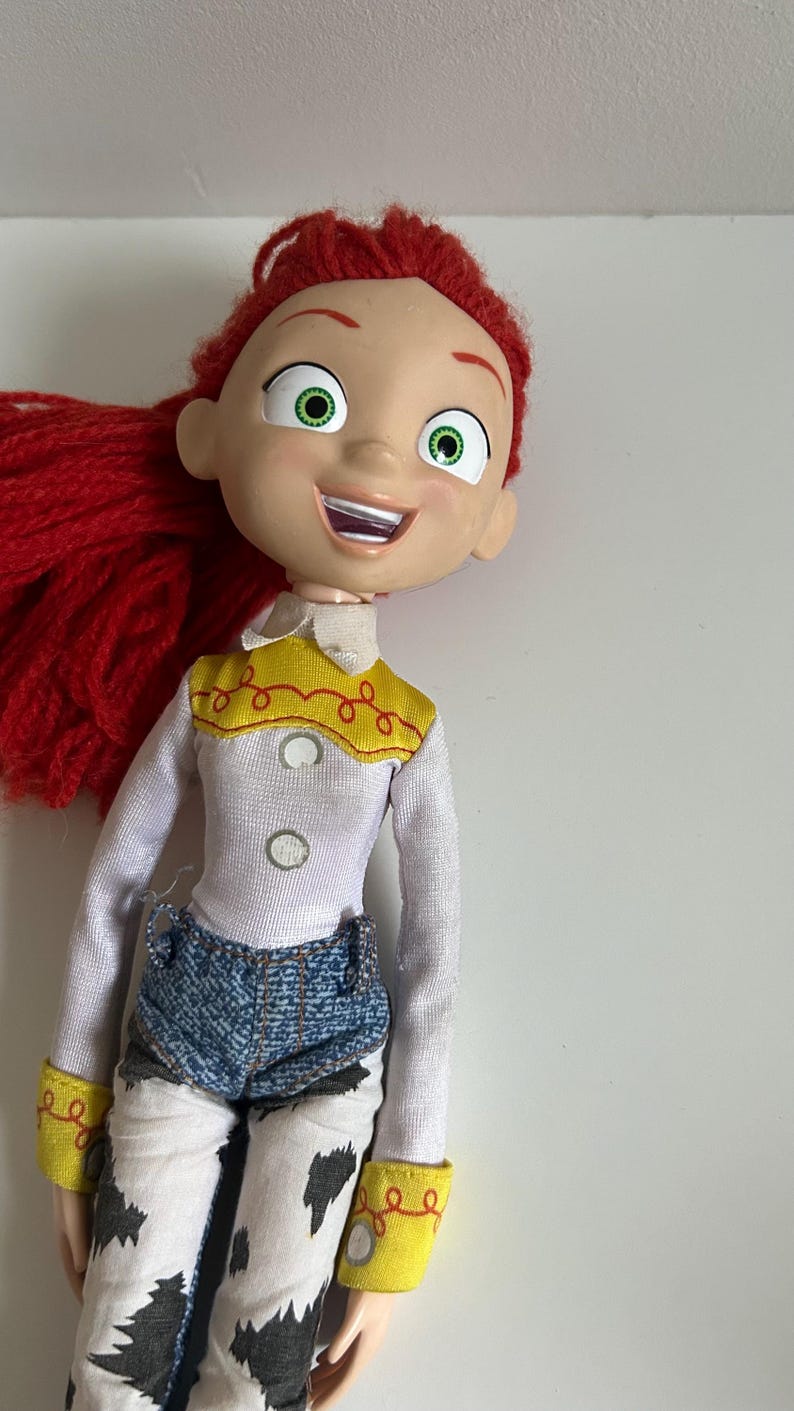 May include: A doll with bright red yarn hair, large green eyes, and an open-mouthed smile. The doll wears a white long-sleeved shirt with a yellow yoke featuring red stitching, two white buttons, blue denim shorts, and white pants with black cow-print spots.