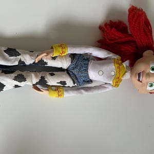 May include: A doll with bright red yarn hair, large green eyes, and an open-mouthed expression. The doll wears a white long-sleeved shirt with yellow trim, a blue textured vest, and white flared pants featuring a black cow-print pattern.
