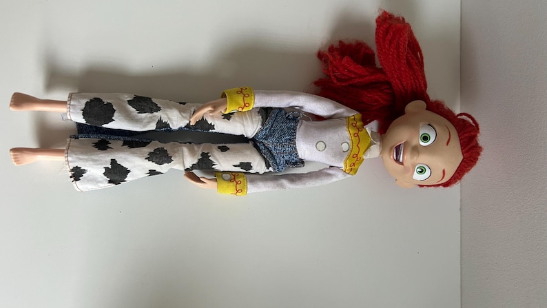 May include: A doll with bright red yarn hair, large green eyes, and an open-mouthed expression. The doll wears a white long-sleeved shirt with yellow trim, a blue textured vest, and white flared pants featuring a black cow-print pattern.