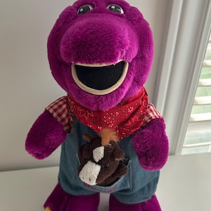 May include: A purple plush dinosaur toy wearing blue denim overalls, a red and white checkered shirt, and a red bandana. A small brown horse plush with a white blaze on its face is tucked into the front pocket of the overalls. The dinosaur has yellow feet.