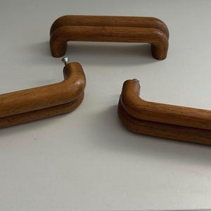 May include: Three medium-brown wooden cabinet handles with a smooth, rounded, double-bar design. Each handle features a curved, arch-like shape with visible wood grain texture. Two of the handles have silver-coloured metal screws attached to ends.