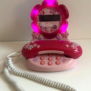 May include: A pink Barbie landline telephone with a flower-shaped base and handset. The device features a digital display screen, a coiled white cord, and flower-shaped buttons. The top flower frame glows with a bright pink light when powered on.