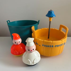 May include: A selection of vintage Fisher-Price toys. Features a teal bucket, a yellow tub with a blue shower head, and two bobble-head figures in red and white outfits. The Fisher-Price logo is printed on the yellow tub.