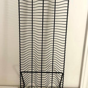 May include: A tall, black metal wire storage rack featuring a series of horizontal, wavy rungs stacked vertically. The structure is supported by a sturdy base with four feet and topped with five small, spherical finials at the top of each wire.