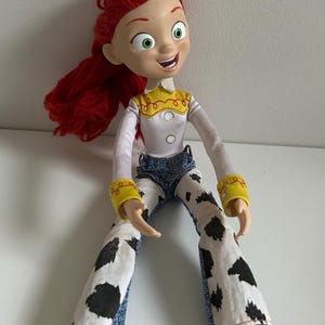 May include: A plastic doll with bright red yarn hair, large green eyes, and a smiling expression. The doll wears a white long-sleeved shirt with a yellow collar, yellow cuffs, and blue denim pants featuring a white and black cow-print pattern.
