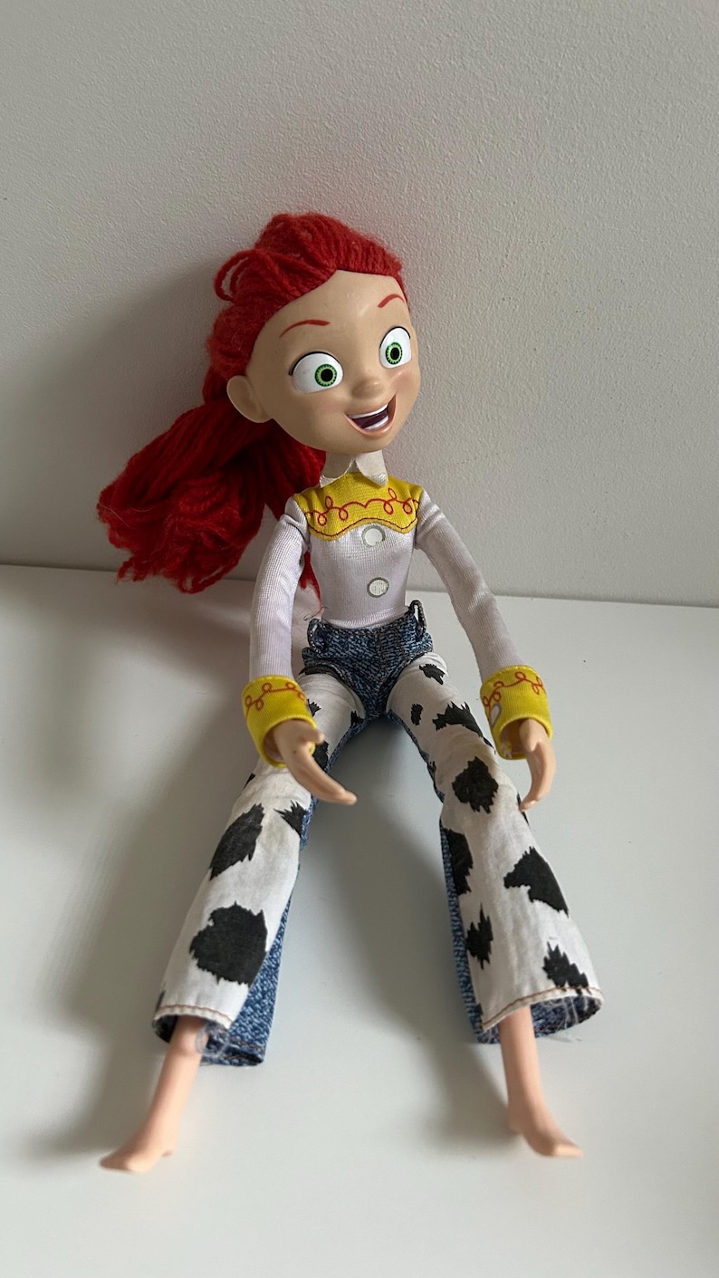 May include: A plastic doll with bright red yarn hair, large green eyes, and a smiling expression. The doll wears a white long-sleeved shirt with a yellow collar, yellow cuffs, and blue denim pants featuring a white and black cow-print pattern.