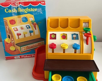 Puede incluir: Un juguete de caja registradora Fisher-Price vintage. La caja registradora de plástico amarillo presenta botones coloridos, un cajón con monedas y un tobogán rojo. La caja, con las palabras "Cash Register", es visible en el fondo.