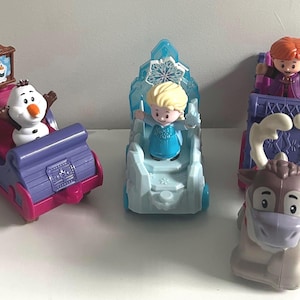 May include: Three plastic toy figures from the Frozen franchise. One is a white snowman in a purple and pink sleigh, one is a blonde character in a light blue ice chariot, and one is a brown reindeer pulling a purple carriage with a red-haired figure.