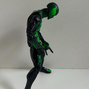 May include: A posable action figure with a black and metallic green color scheme. The figure features a sleek, humanoid design with bright green accents on the head, torso, and limbs, and is posed in a standing position with articulated joints.