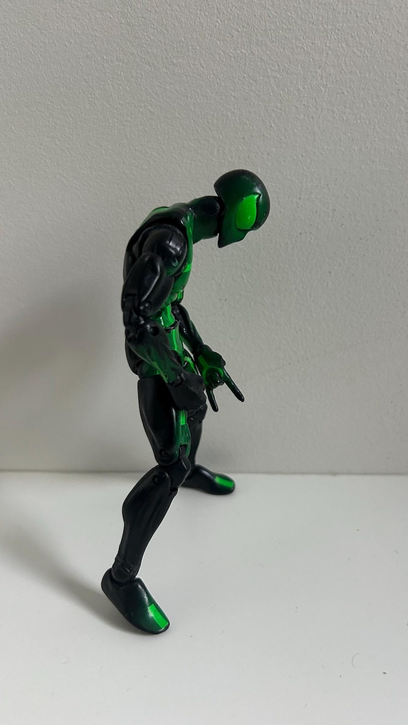 May include: A posable action figure with a black and metallic green color scheme. The figure features a sleek, humanoid design with bright green accents on the head, torso, and limbs, and is posed in a standing position with articulated joints.