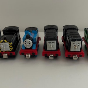 May include: A collection of six small toy train engines arranged in a row. The set includes two black engines with yellow hazard stripes, a blue engine with a face, two black engines with grey faces, and one green engine with a face.