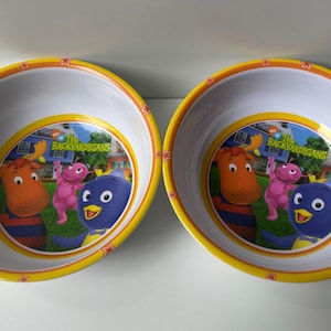 May include: Two round white bowls with yellow and orange rims featuring a graphic print of The Backyardigans characters Tyrone, Uniqua, and Pablo on a green lawn. The text The Backyardigans is printed in green and yellow above the characters.