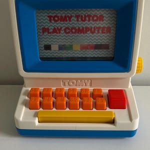 May include: A white and blue Tomy Tutor Play Computer toy with a screen displaying the text TOMY TUTOR PLAY COMPUTER and a colour bar. The device features a row of orange keys, a large red square button, and a long yellow space bar on the front.