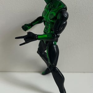 May include: A posable action figure featuring a black and metallic green suit with a full-face mask and large, bright green eye lenses. The figure is captured in a dynamic stance with one hand extended forward in a web-shooting gesture.