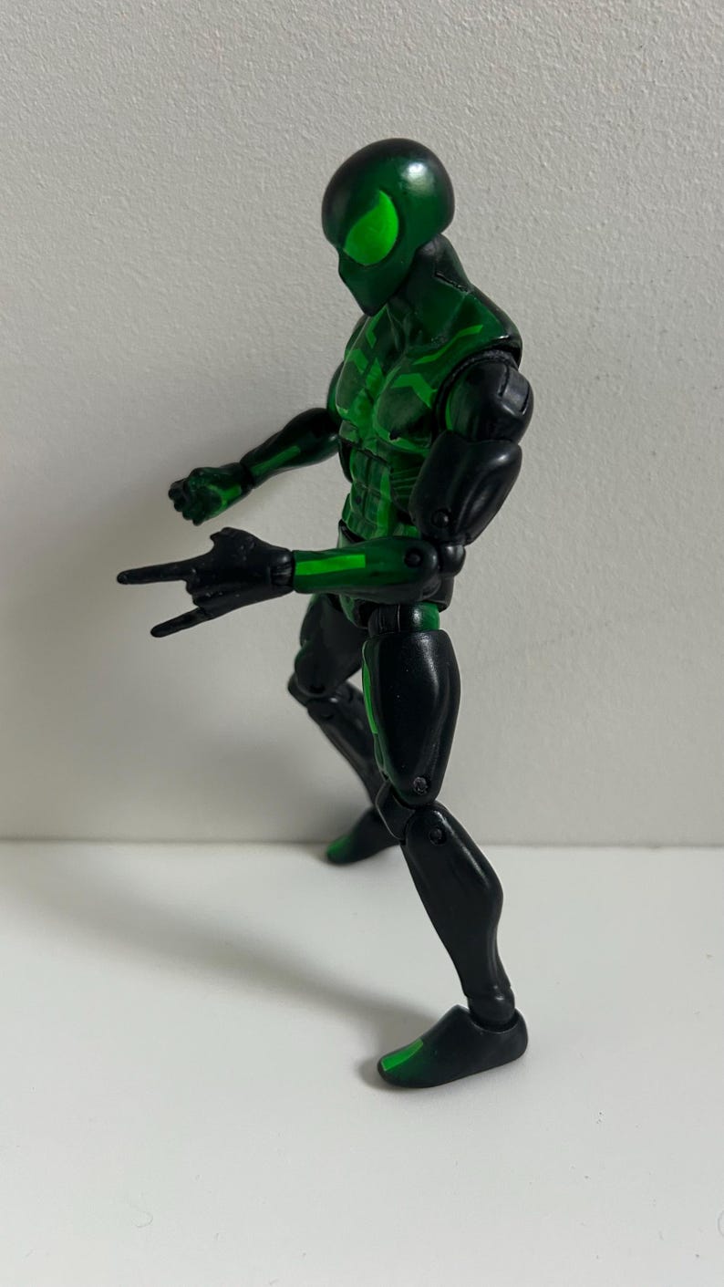 May include: A posable action figure featuring a black and metallic green suit with a full-face mask and large, bright green eye lenses. The figure is captured in a dynamic stance with one hand extended forward in a web-shooting gesture.