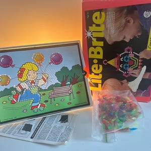 Peut inclure: Un jeu Lite-Brite avec un écran lumineux représentant un patineur à roulettes de dessin animé tenant des ballons. La boîte indique "Lite-Brite" et "Créez des images colorées et lumineuses !" Un sac de chevilles colorées et des instructions sont également inclus.