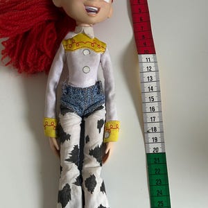 May include: A Jessie doll with red yarn hair, green eyes, and a white long-sleeve shirt with a yellow yoke. The doll wears blue denim shorts over white pants with a black cow-print pattern. A measuring tape shows the doll is 12 inches tall.