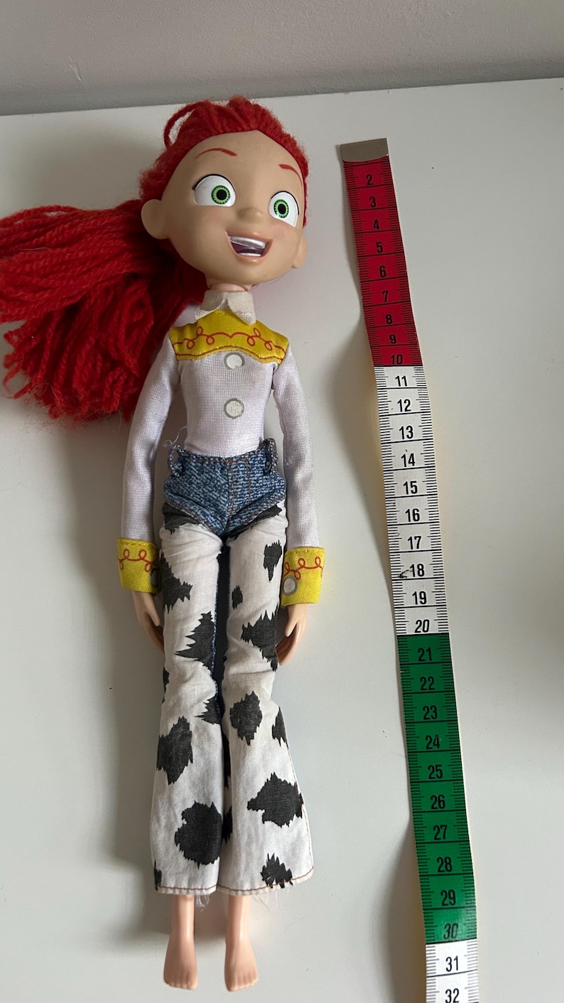 May include: A Jessie doll with red yarn hair, green eyes, and a white long-sleeve shirt with a yellow yoke. The doll wears blue denim shorts over white pants with a black cow-print pattern. A measuring tape shows the doll is 12 inches tall.