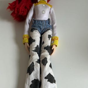 May include: A doll with bright red yarn hair, green eyes, and an open mouth expression. The doll wears a white long-sleeved shirt with a yellow collar, blue denim-style shorts, and white pants featuring a black cow-print pattern with yellow cuffs.