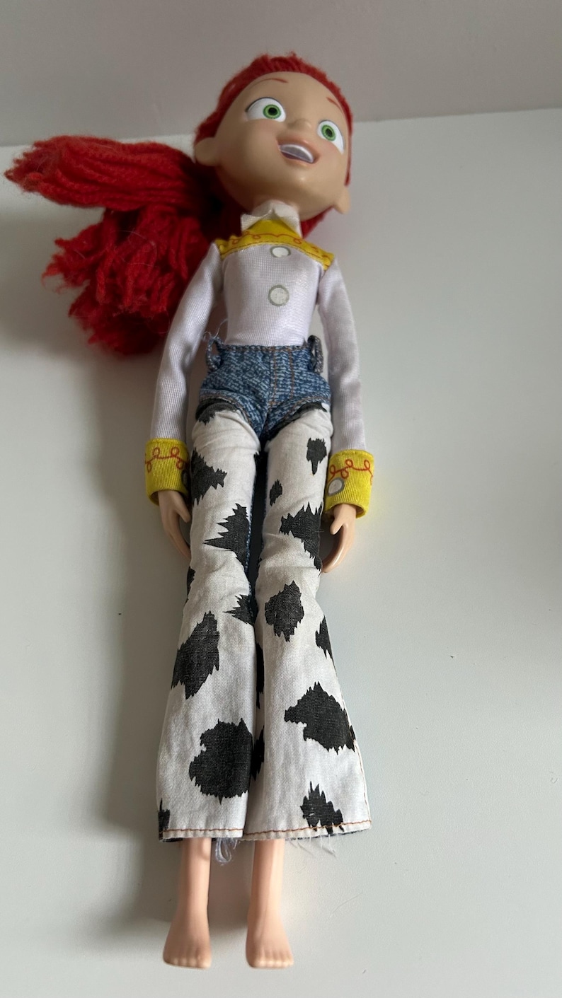 May include: A doll with bright red yarn hair, green eyes, and an open mouth expression. The doll wears a white long-sleeved shirt with a yellow collar, blue denim-style shorts, and white pants featuring a black cow-print pattern with yellow cuffs.