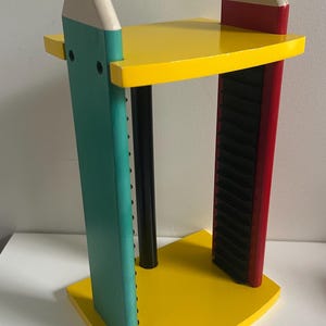 May include: A media storage rack featuring two vertical supports shaped like oversized pencils in teal and red. The unit has yellow top and bottom shelves, a central black cylindrical support, and black grooved side panels for holding media cases.