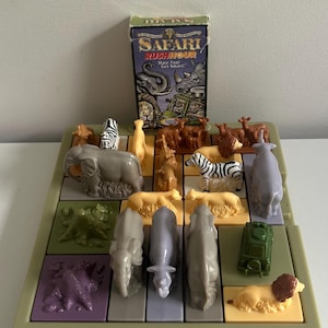 May include: A Safari Rush Hour board game by Binary Arts featuring a light green plastic grid base with various molded plastic animal figures including elephants, zebras, lions, and a green vehicle. The game box stands upright behind the board.
