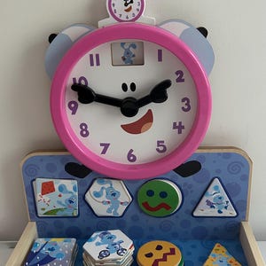 Peut inclure: Un jouet horloge d'apprentissage en bois Blue's Clues avec un cadre rose, des aiguilles noires et un cadran blanc affichant les chiffres de 1 à 12. Sous l'horloge se trouve un plateau bleu avec des formes géométriques et Melissa & Doug.
