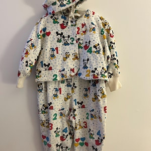 May include: A white baby outfit with a hooded jacket and trousers. The outfit is patterned with colourful images of Mickey Mouse, Donald Duck, and other Disney characters, along with numbers and hearts. The cuffs and hood lining are white.