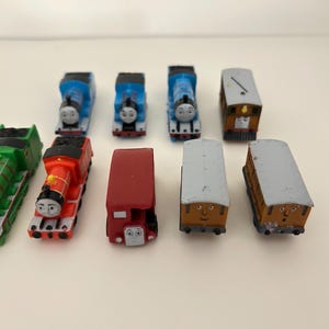 May include: A selection of toy train engines and carriages in various colours. The set includes blue, red, green, and brown engines and carriages. The toy trains are arranged on a white surface.