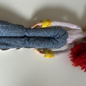 May include: A doll with bright red yarn hair, wearing a white long-sleeved shirt with yellow cuffs featuring red swirl patterns, and blue denim-style pants with a black and white cow-print pattern on the lower legs, shown from a side angle.