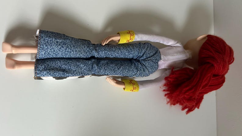 May include: A doll with bright red yarn hair, wearing a white long-sleeved shirt with yellow cuffs featuring red swirl patterns, and blue denim-style pants with a black and white cow-print pattern on the lower legs, shown from a side angle.