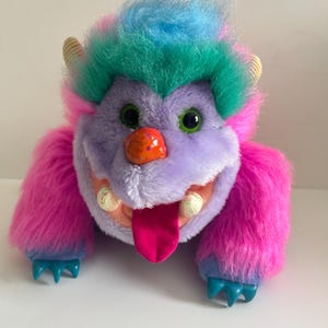 Peut inclure: Une peluche monstre avec un visage violet, des bras en fourrure rose et des pieds bleus. Le monstre a des yeux verts, un nez orange et une langue rose. Ses cheveux sont un mélange de bleu et de vert.