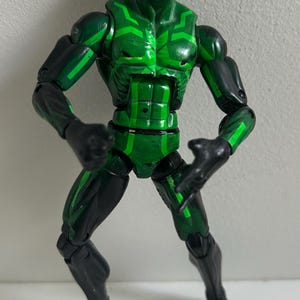 May include: A posable action figure featuring a black and green color scheme with bright green glowing-style eyes. The figure has a muscular build with articulated joints at the shoulders, elbows, hips, and knees, displaying a geometric pattern.