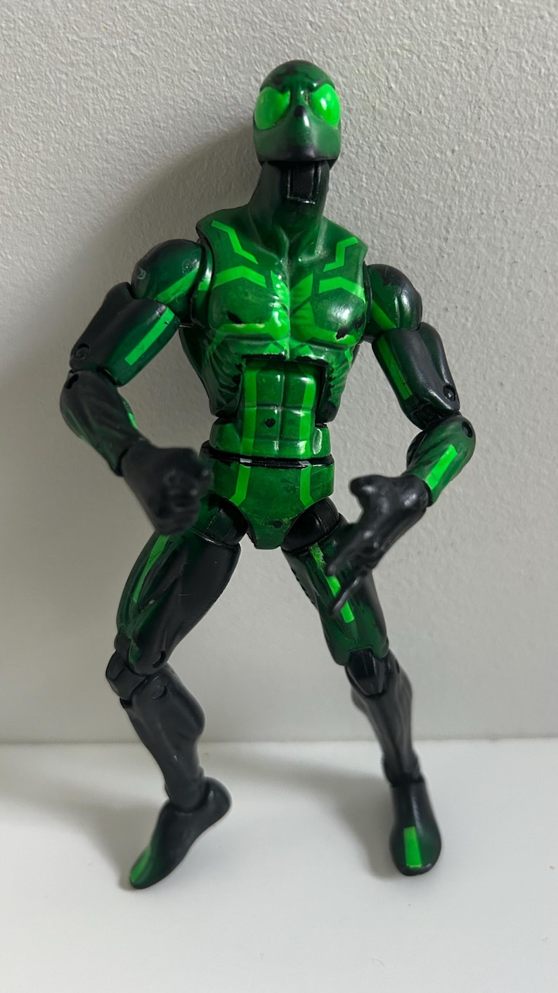 May include: A posable action figure featuring a black and green color scheme with bright green glowing-style eyes. The figure has a muscular build with articulated joints at the shoulders, elbows, hips, and knees, displaying a geometric pattern.