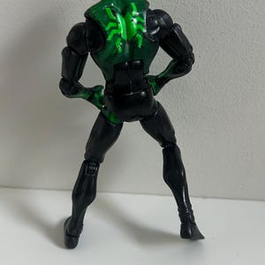 May include: A posable action figure featuring a black and green color scheme. The figure has a solid black head, arms, and legs, with a green torso section displaying a bright green spider-like emblem on the back. The figure stands upright.