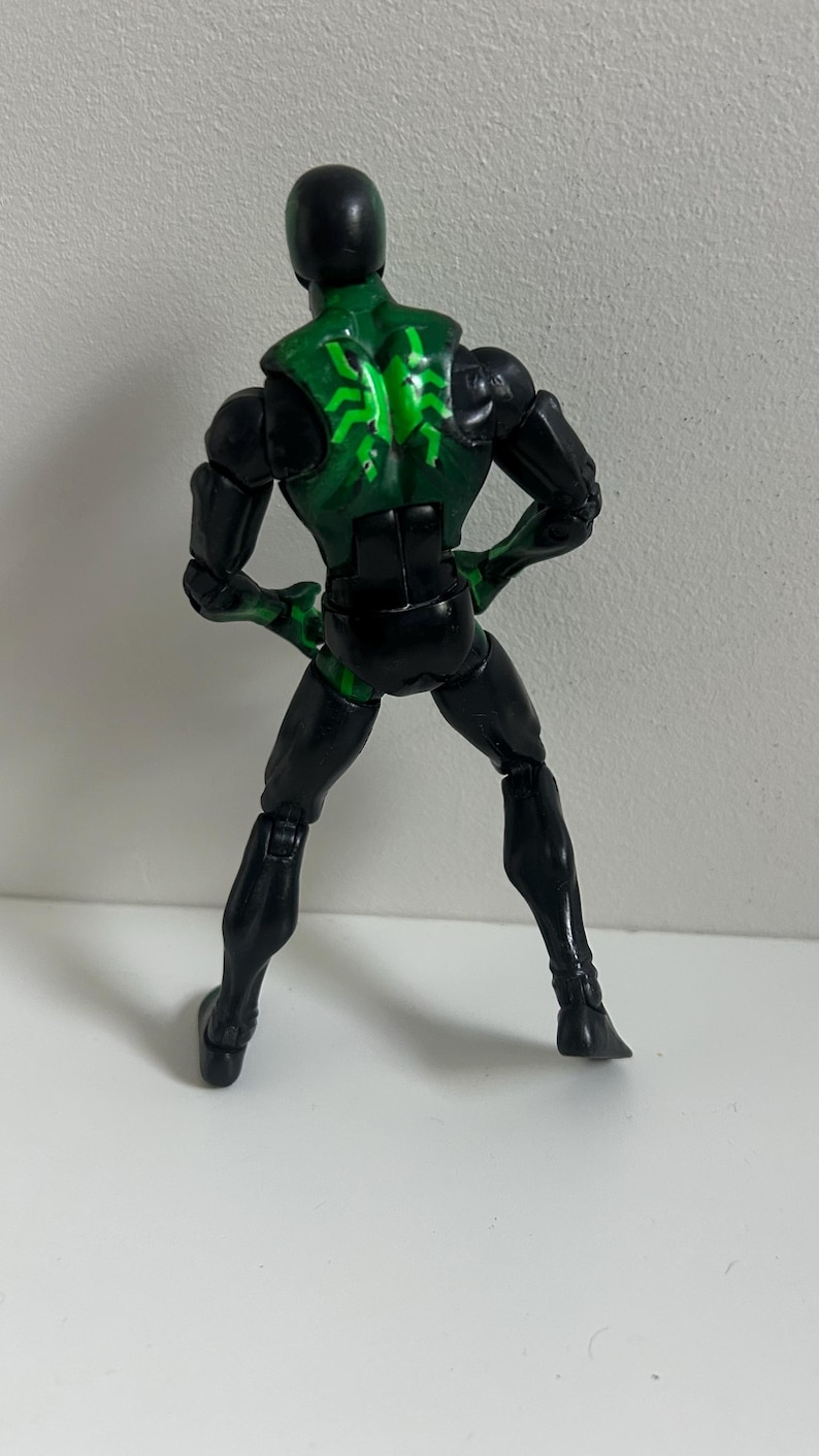 May include: A posable action figure featuring a black and green color scheme. The figure has a solid black head, arms, and legs, with a green torso section displaying a bright green spider-like emblem on the back. The figure stands upright.