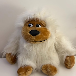 Peut inclure: Une peluche de yéti avec une fourrure blanche et un visage, des mains et des pieds beiges. Le yéti a une expression amicale avec des yeux noirs et un nez noir. Le jouet est posé sur une surface blanche.