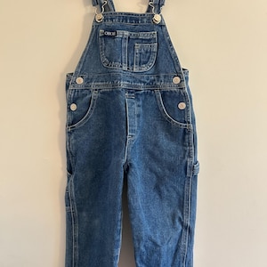 May include: Blue denim overalls with adjustable straps and metal hardware. The overalls have a bib pocket, side pockets, and button closures. The brand name "Cherokee" is visible on the bib. A classic, durable garment for children.