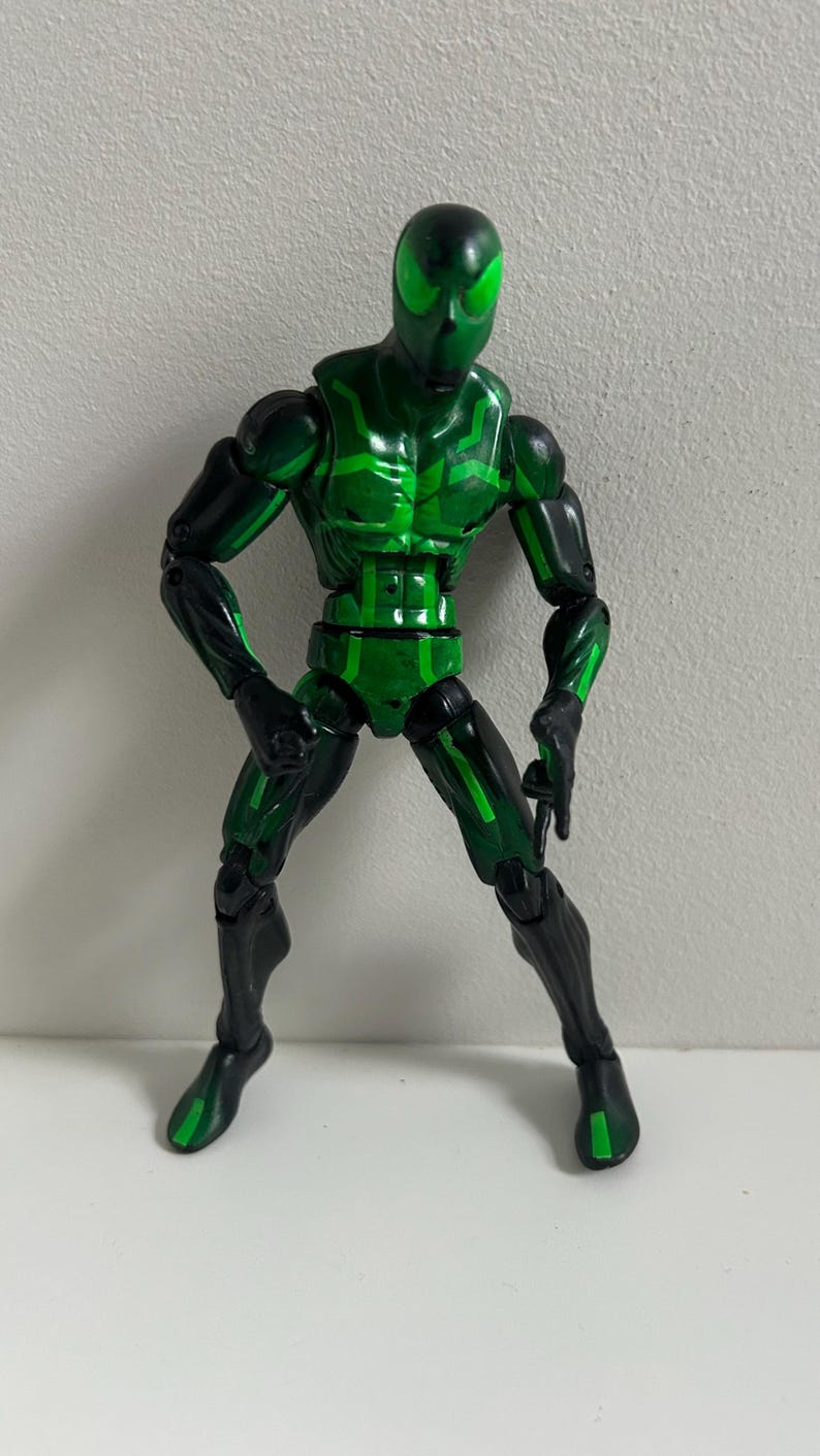 May include: A posable action figure featuring a black and bright green color scheme. The figure has a smooth, mask-like head with large, vibrant green eyes, a muscular torso with geometric green patterns, and articulated limbs in black and green.