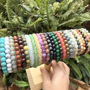 May include: A display of beaded bracelets in various colors, including turquoise, brown, green, and purple. The bracelets are made of different types of stones and are arranged on a wooden stand. The background features green foliage.