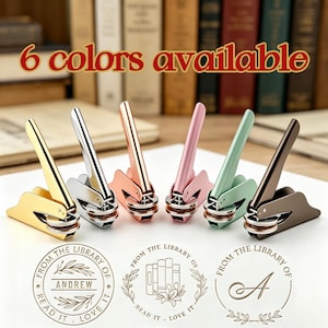 Può includere: Sei sigilli di biblioteca in metallo color oro, argento, oro rosa, rosa, verde menta e bronzo. Ogni sigillo ha un manico e una base circolare. Il testo "6 colori disponibili" è visualizzato sopra i sigilli. Sotto ci sono tre esempi di design di timbri.