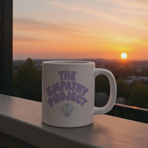 May include: White ceramic mug with the text "THE EMPATHY PROJECT" in purple and light blue. The mug features a handle and a design of a heart and hands. The background shows a sunset.