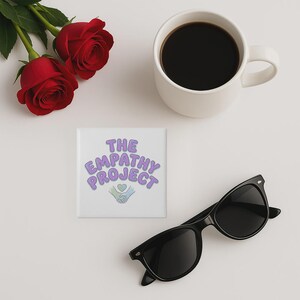 May include: A square white coaster with the text "THE EMPATHY PROJECT" in purple and blue lettering, with a heart and hands graphic. Also, a pair of black sunglasses, a white mug of coffee, and two red roses.