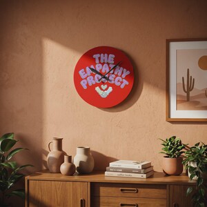 May include: A red wall clock with the text "The Empathy Project" in light purple and a graphic of two hands shaking with a heart. The clock is round and has black clock hands. The clock is hanging on a peach-colored wall.
