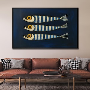 Può includere: Opera d'arte incorniciata con tre pesci stilizzati. Ogni pesce ha un corpo color crema con strisce blu, occhi gialli e pinna caudale bianca. Lo sfondo è blu scuro e la cornice nera.