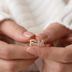 Puede incluir: Un anillo de tono dorado que presenta dos bandas separadas, cada una rematada con una letra mayúscula, F y M, respectivamente. Ambas letras están incrustadas con pequeñas piedras claras y brillantes que captan la luz, creando un aspecto refinado.