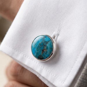 Op de afbeelding: Close-up van een zilveren manchetknop met een ronde, turquoise steen. De steen heeft een gemarmerd patroon met blauwe en bruine tinten. De manchetknop is bevestigd aan een wit overhemd.