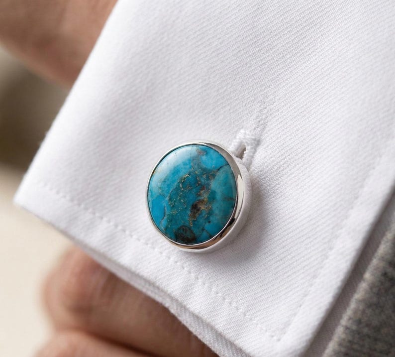 Op de afbeelding: Close-up van een zilveren manchetknop met een ronde, turquoise steen. De steen heeft een gemarmerd patroon met blauwe en bruine tinten. De manchetknop is bevestigd aan een wit overhemd.